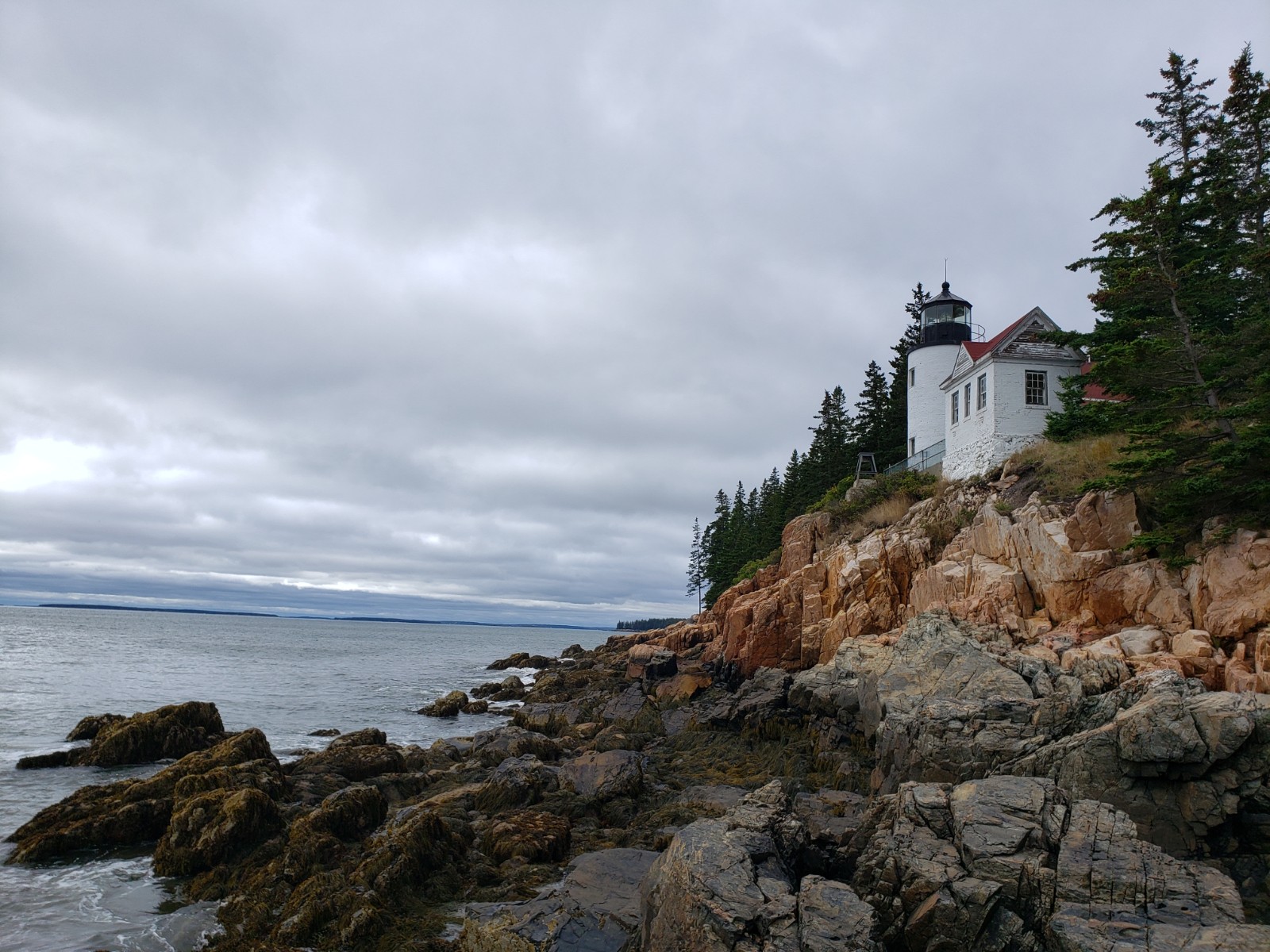 A gray sky and sea surround a jagged rock coast crowned with a white lighthouse
