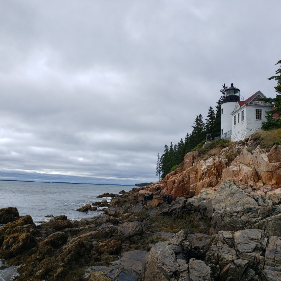 A gray sky and sea surround a jagged rock coast crowned with a white lighthouse