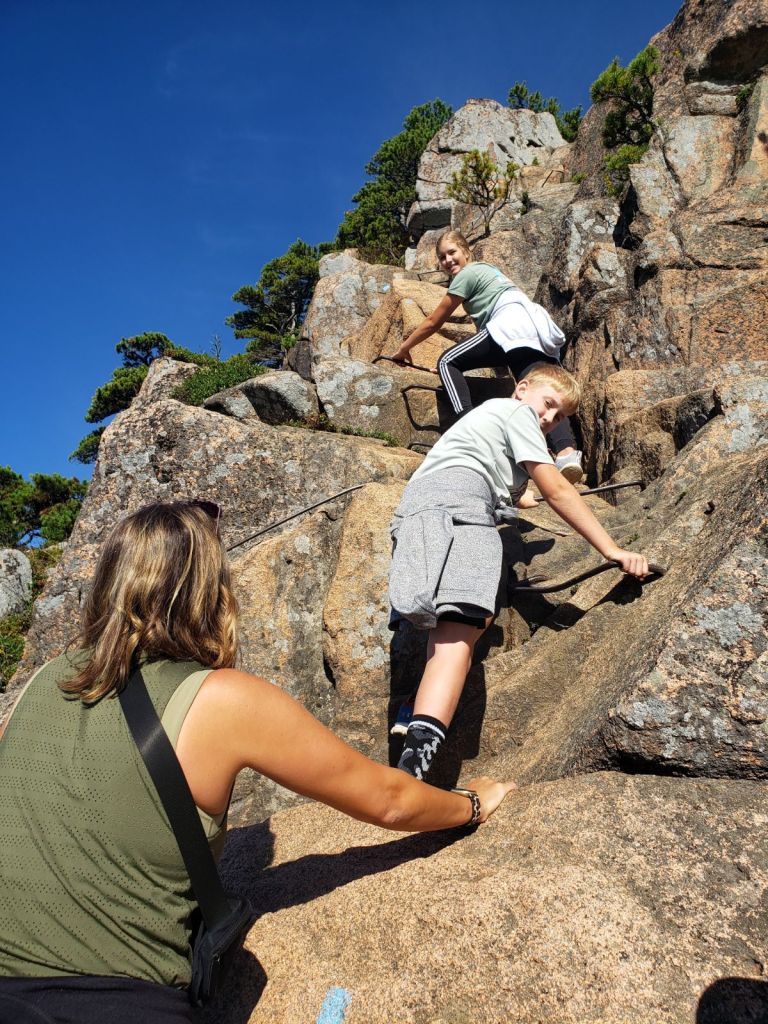 A woman and her two kids scramble up some rocks with iron rungs