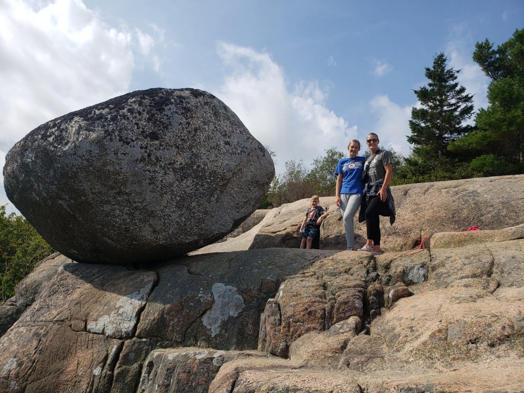 A boy, girl, and mother stand beside a large bolder that balances over a cliff