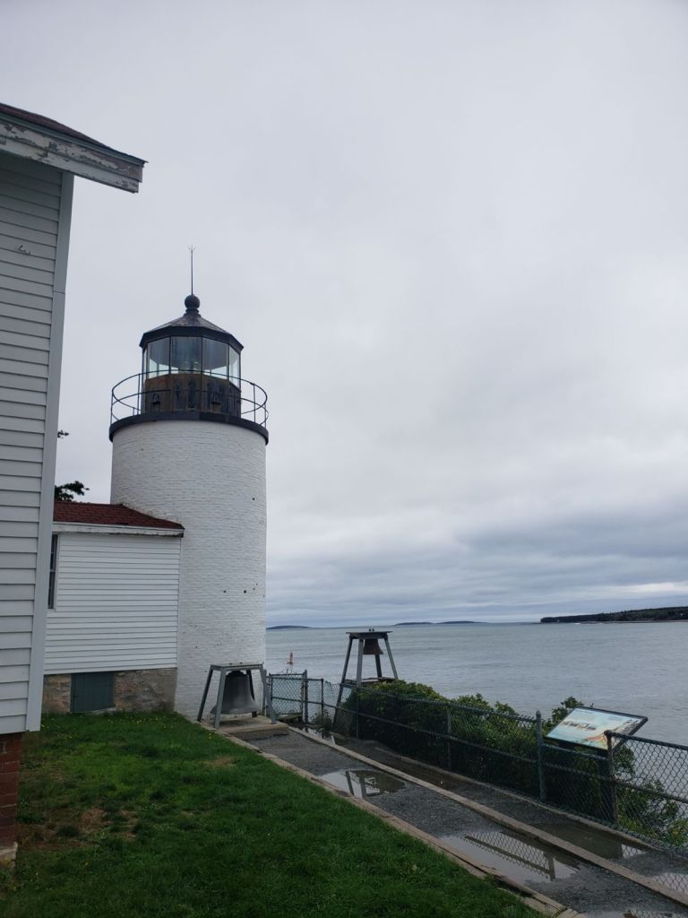 A white lighthouse stands beside a green lawn and a gray sky