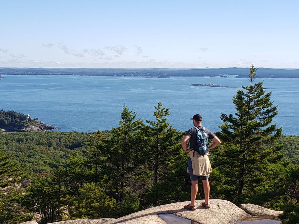 A man in a black ball cap, blue backpack, shorts and sandals stands on a ledge viewing the ocean and green valley before him from high on a mountain