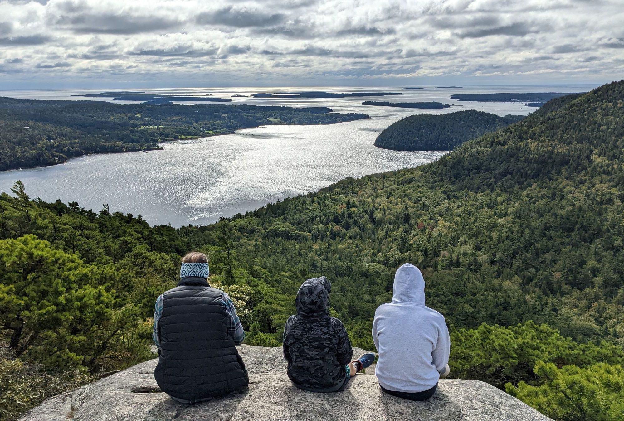 Three people sit with their back to the camera looking at the ocean and valley in front of them