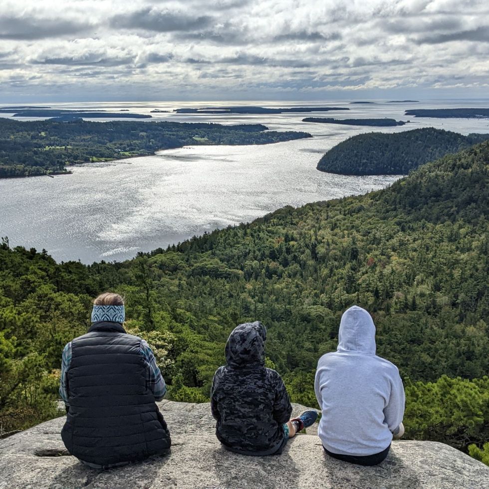 Three people sit with their back to the camera looking at the ocean and valley in front of them