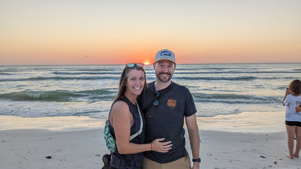 A couple standing on Crescent Beach with an orange sky over the green sea crashing against white sand.