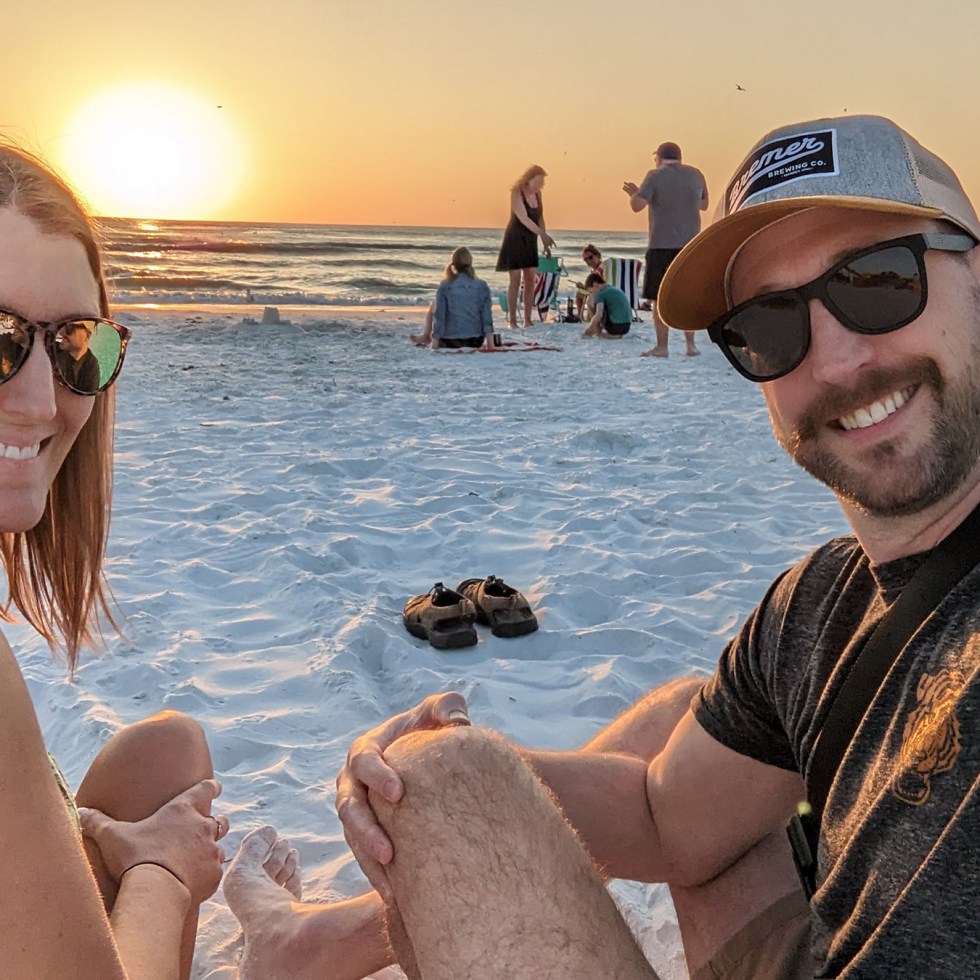 A couple smiling in front of a yellow sun over the ocean crashing against Crescent Beach