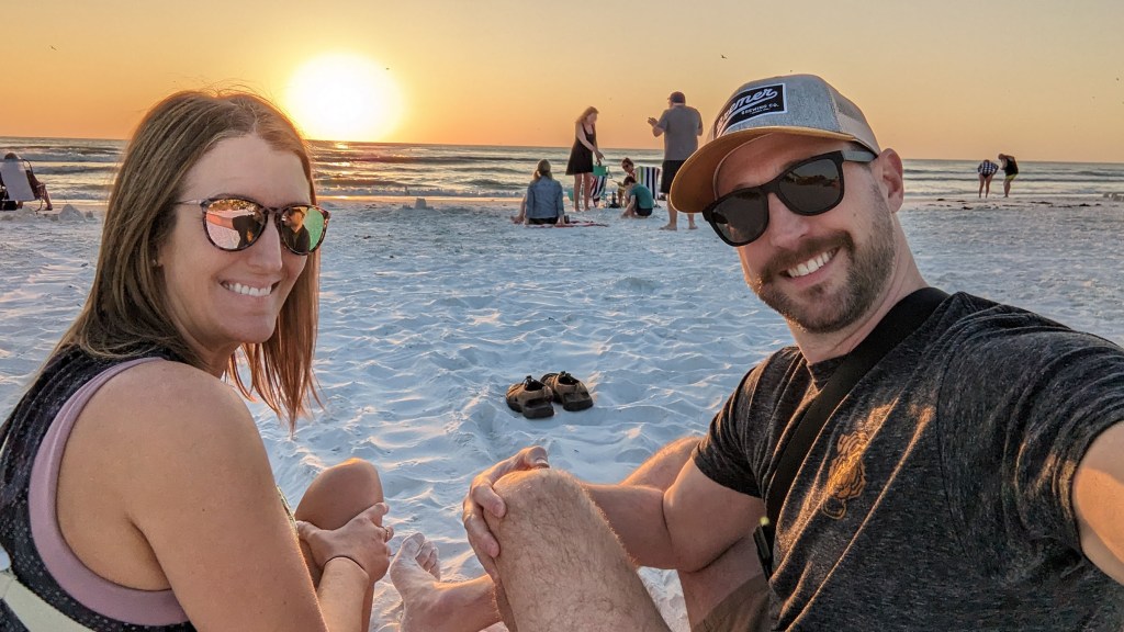A couple smiling in front of a yellow sun over the ocean crashing against Crescent Beach