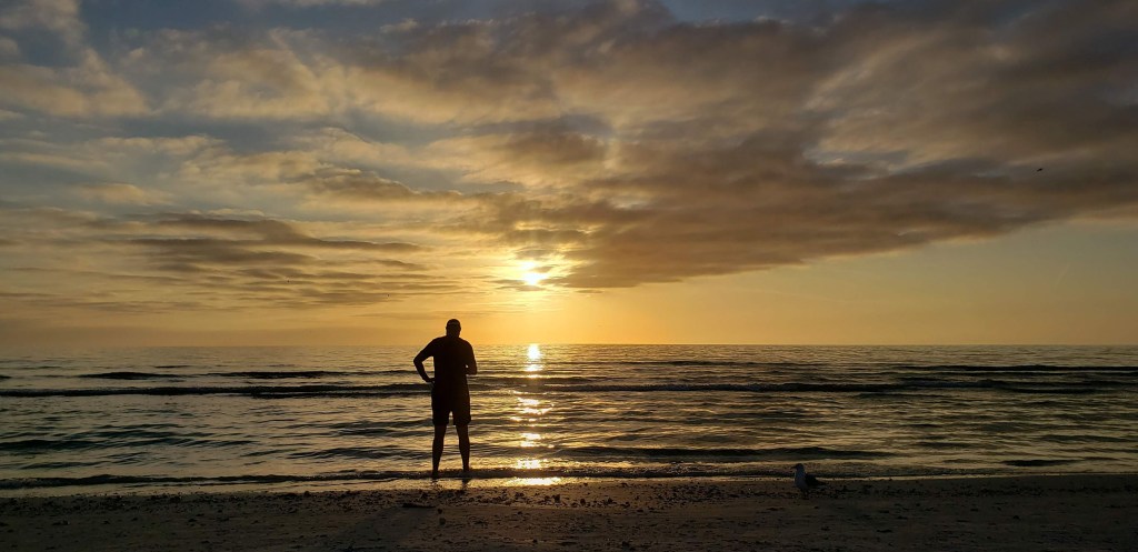 A yellow sun sets over the ocean with a black silhouette of a man standing ankle deep in the ocean