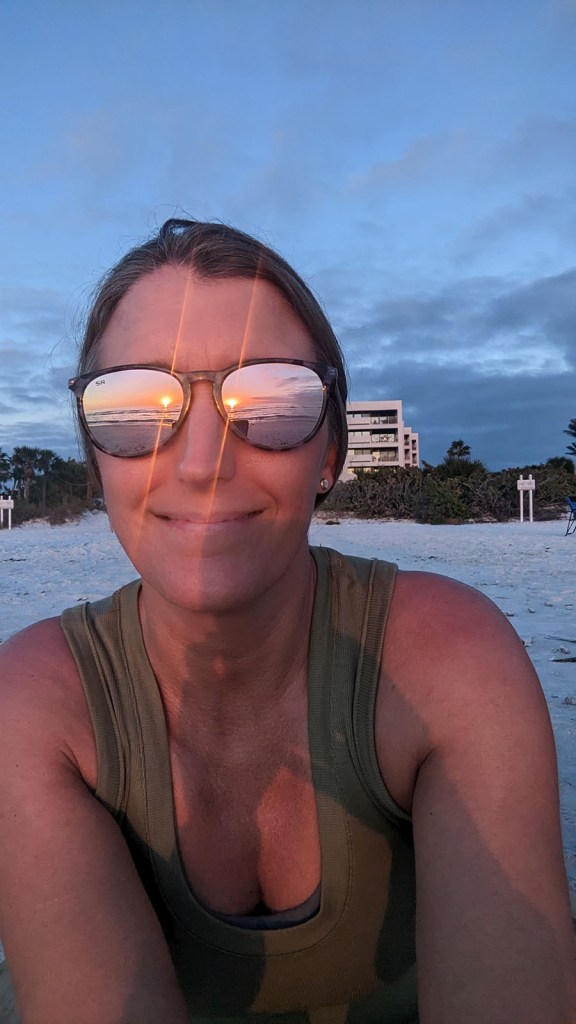 A woman smiles with sunglasses reflecting the sunset