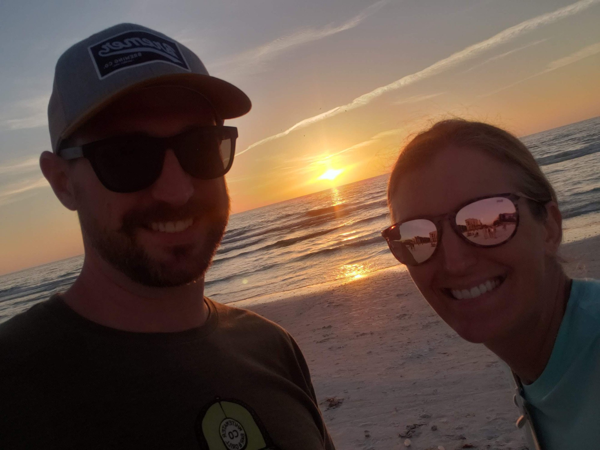 Sunset turns the sky orange behind a smiling couple. The ocean and the beach behind them.