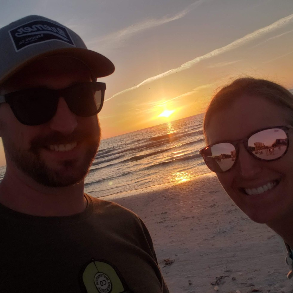 Sunset turns the sky orange behind a smiling couple. The ocean and the beach behind them.