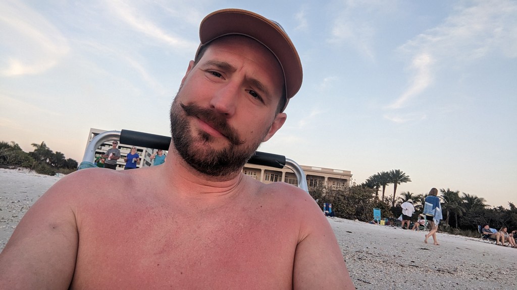 A man with a mustache sitting in a beach chair smiling at the camera