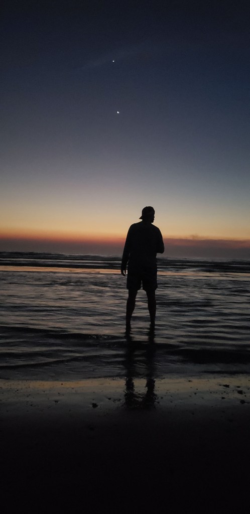 A silhouette of a man in a backwards ballcap standing under two bright planets over the ocean. 