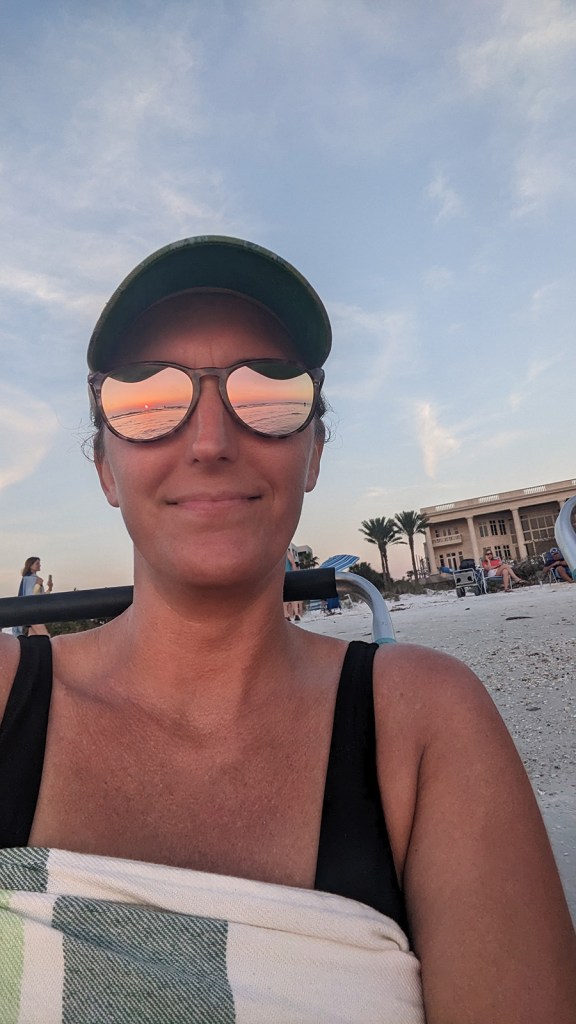 A woman smiling with the sunset reflecting off of her sunglasses.