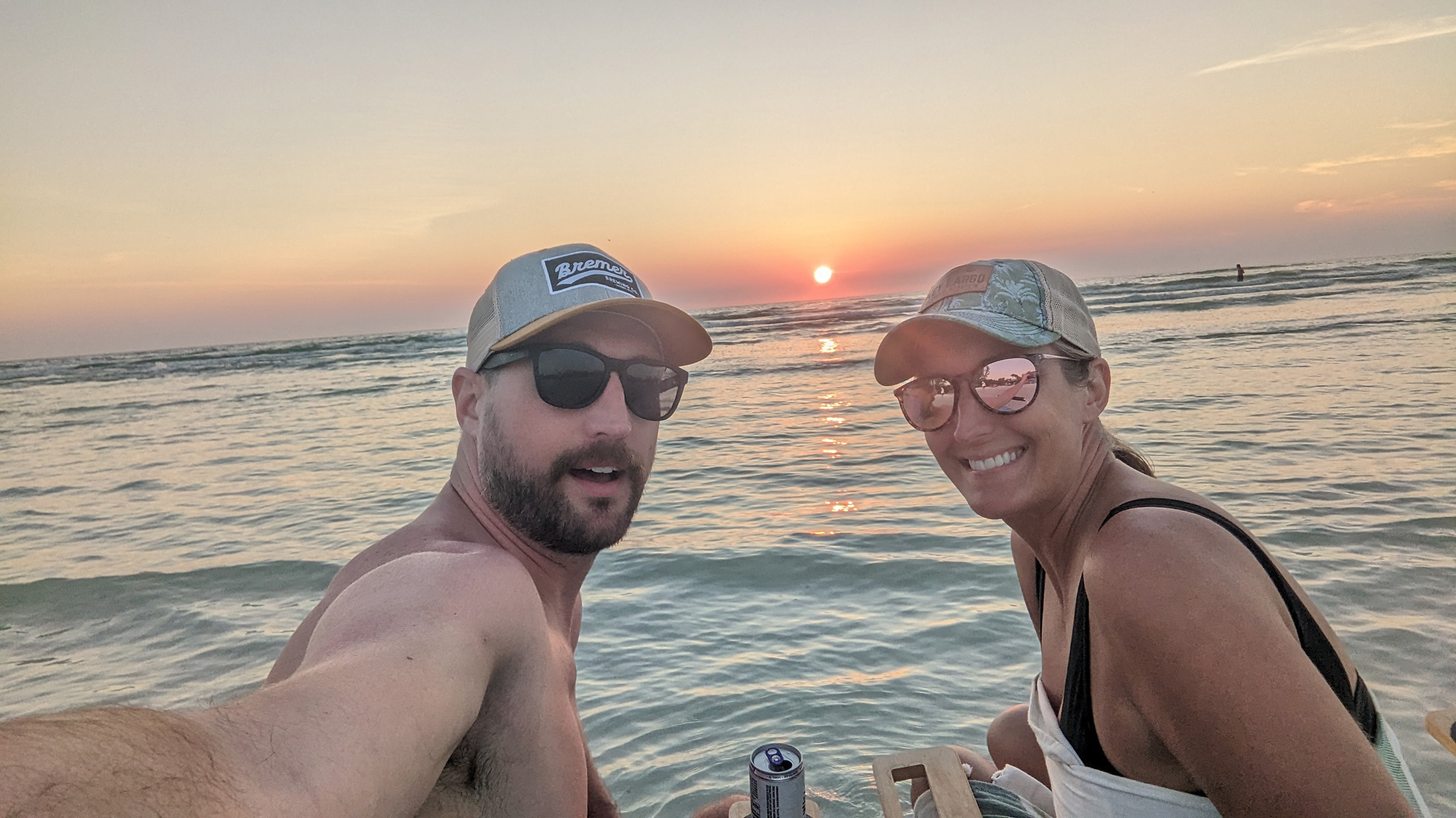 A couple wearing baseball caps smiling with the sun setting in the background over the ocean.