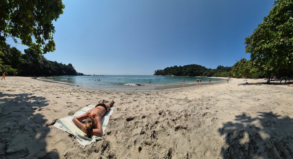 A panoramic picture of a girl laying on a white sand beach with the ocean in the background.