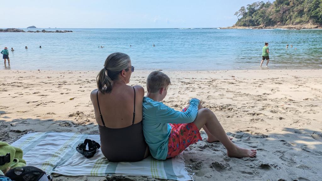 A mother and son sit on a beach towel looking out on the ocean
