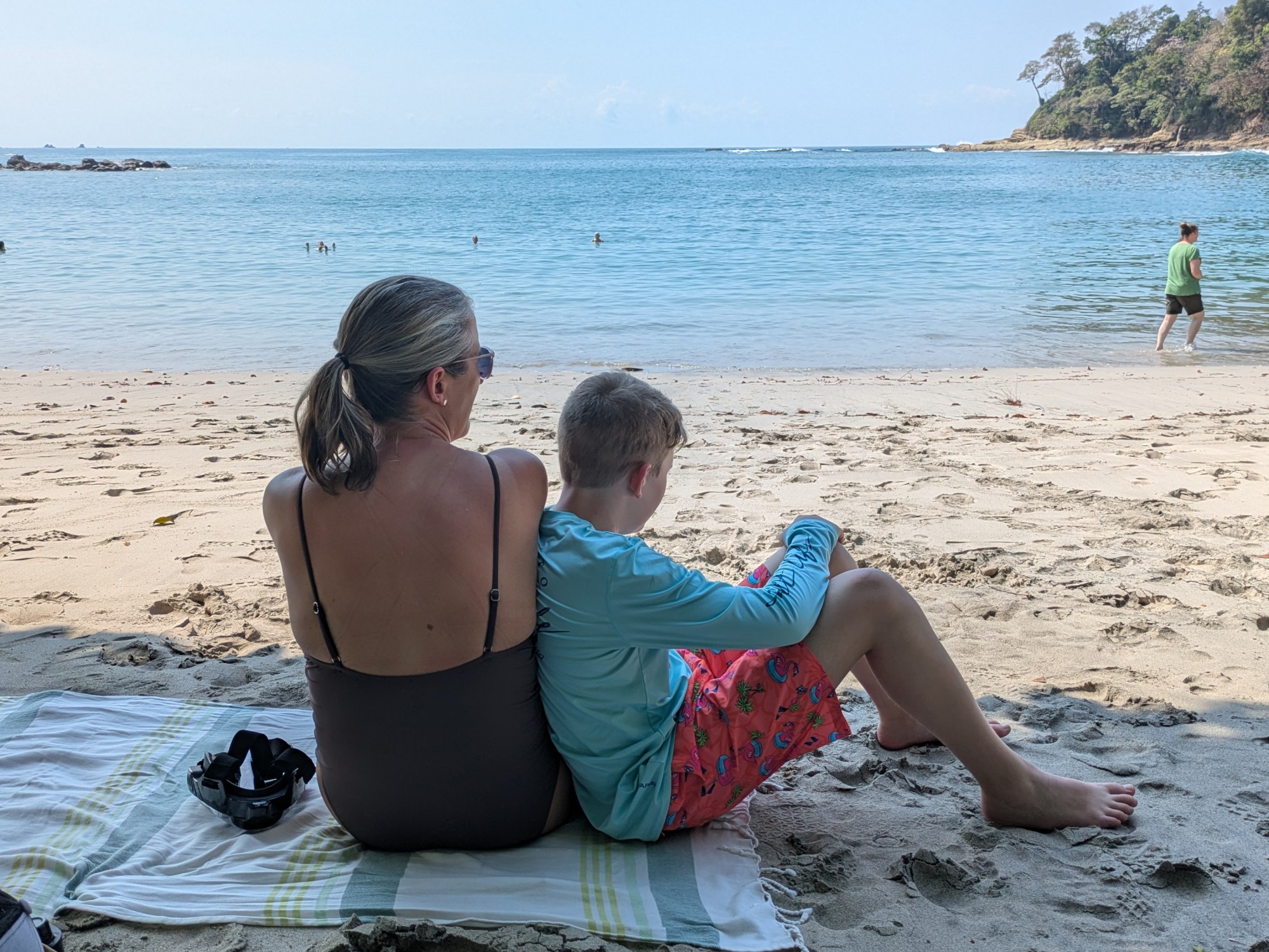 A mother and her son sit on a beach towel looking at the ocean in front of them