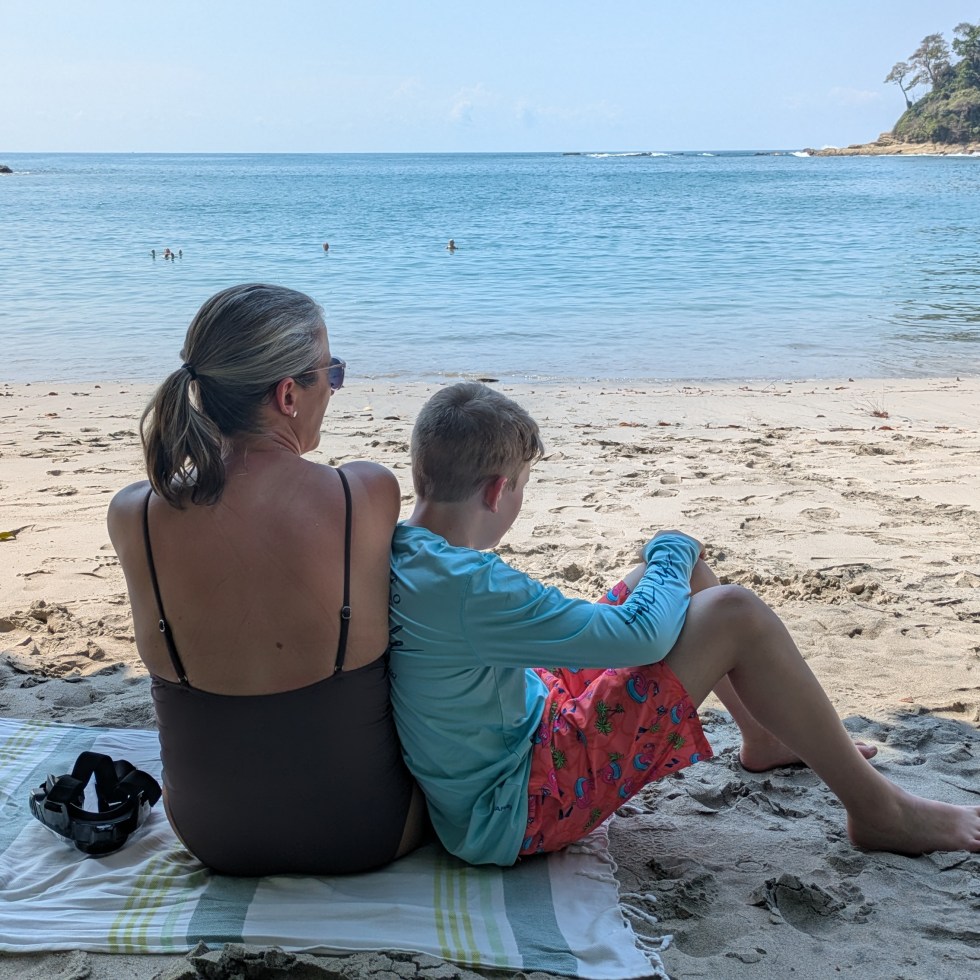 A mother and her son sit on a beach towel looking at the ocean in front of them