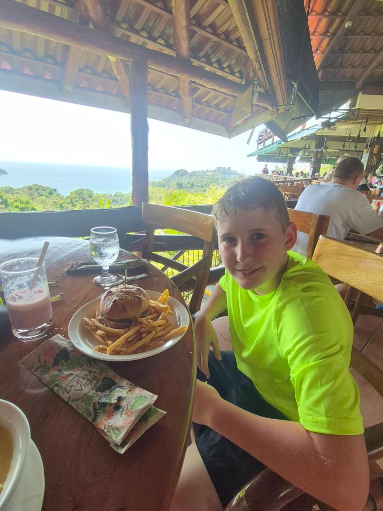 A boy in a bright green shirt sits with a burger at an open air restaurant overlooking the sea