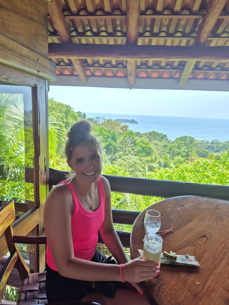 A girl in a bright pink tank top holds a frozen drink with the jungle and sea in the background