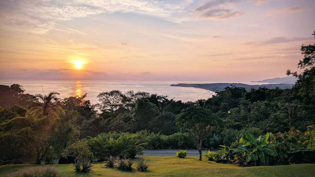 A pink and orange sunset behind a lush hillside of jungle. A beach curls out into the ocean that reflects the sunset.