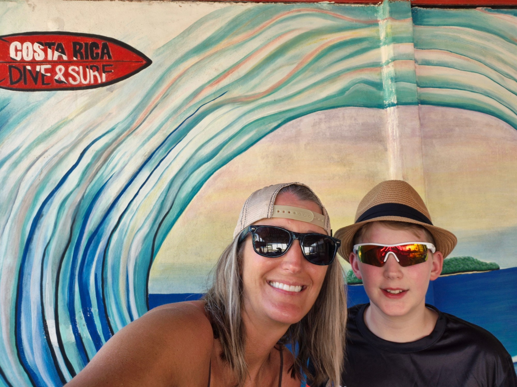A woman and boy in hats and sunglasses smile in front of a mural of a wave.