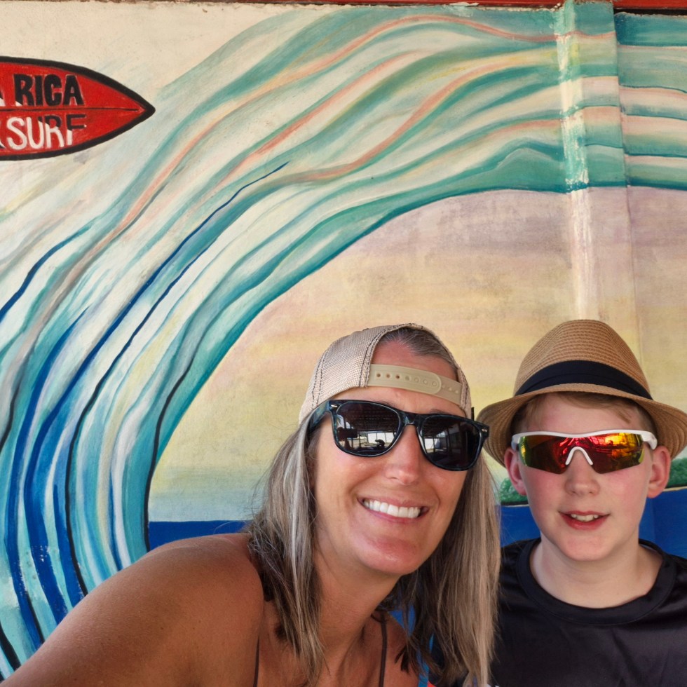 A woman and boy in hats and sunglasses smile in front of a mural of a wave.