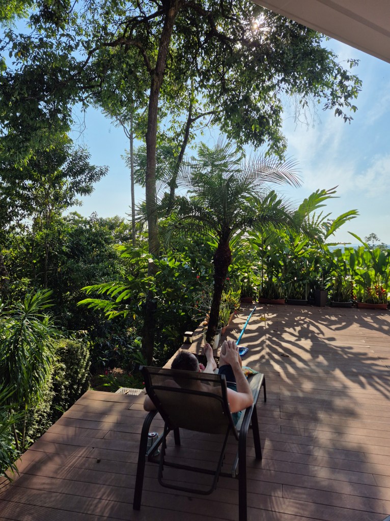 A man sits in at chair in the shade on a deck during a sunny day.