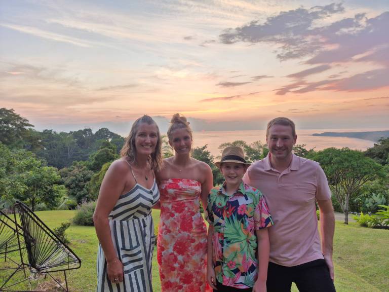 A family of four smiles in front of a grassy ledge and an orange pink sunset over the ocean.