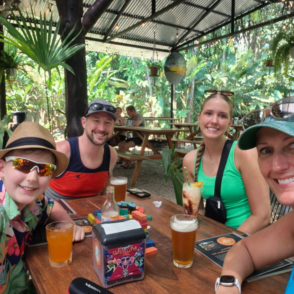 Family in colorful clothing sites at a table surrounded by green plants at a brewery.