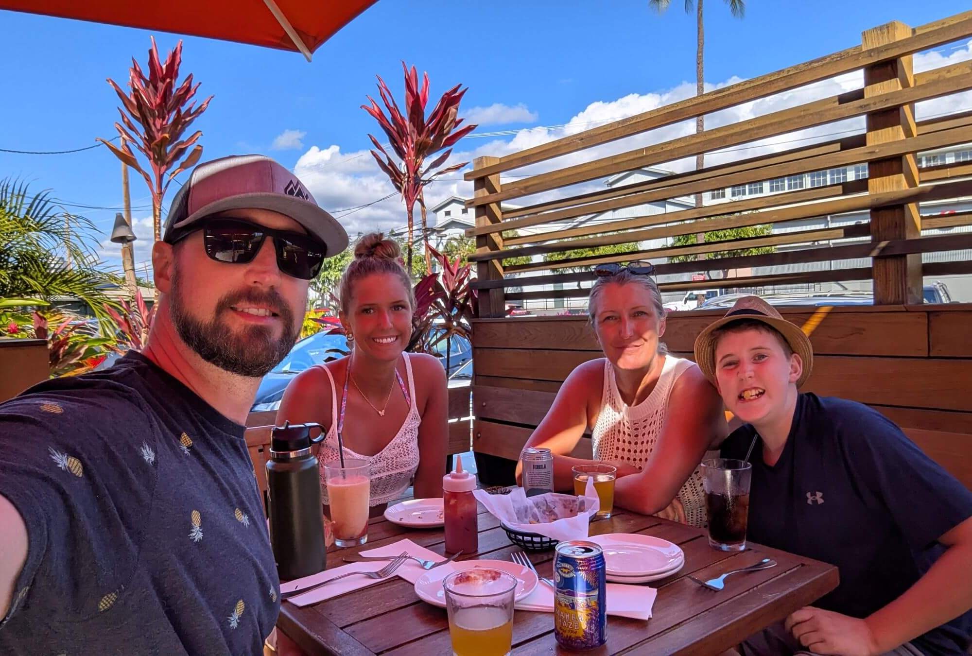 Having Drinks at Aloha Mixed Plate. A family of four sits in the shade a a restaurant table with various drinks on it.