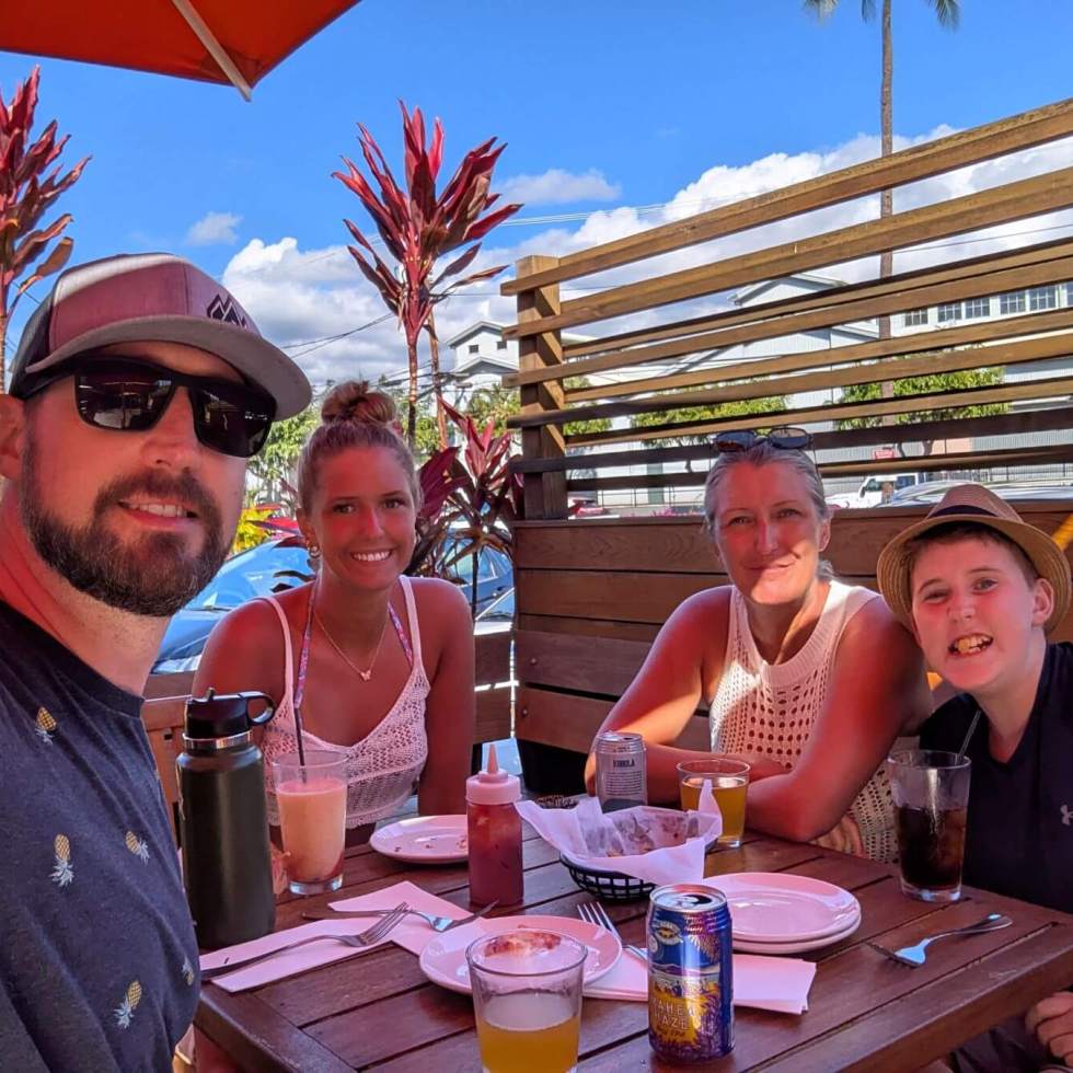 Having Drinks at Aloha Mixed Plate. A family of four sits in the shade a a restaurant table with various drinks on it.
