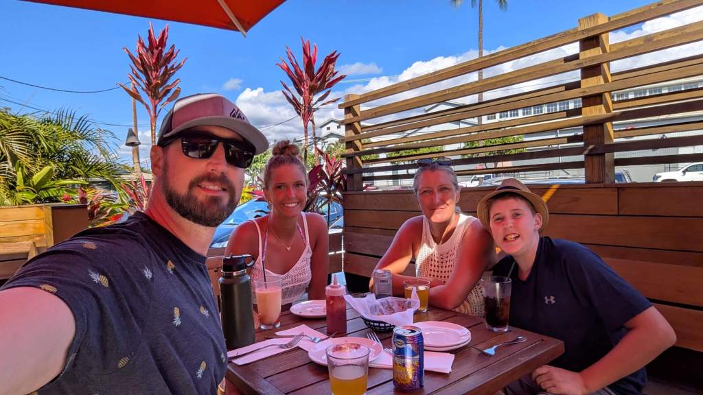 Having Drinks at Aloha Mixed Plate. A family of four sits in the shade a a restaurant table with various drinks on it.