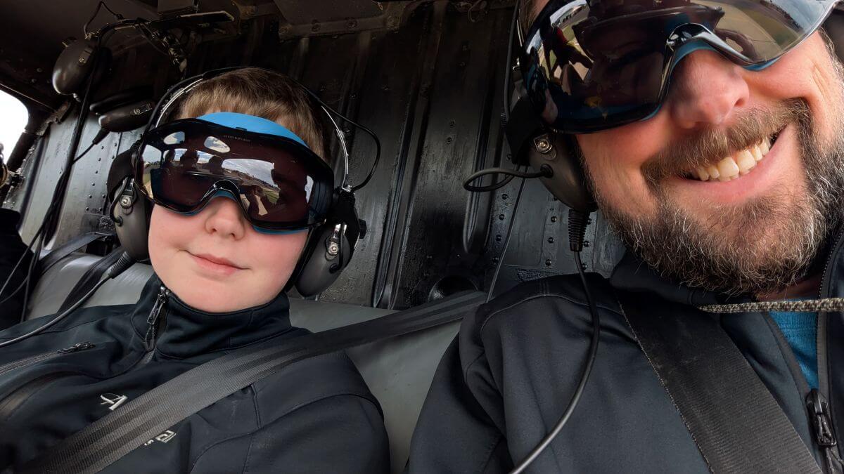 A father takes a selfie with his son in the backseat of a helicopter wearing goggles and large headphones