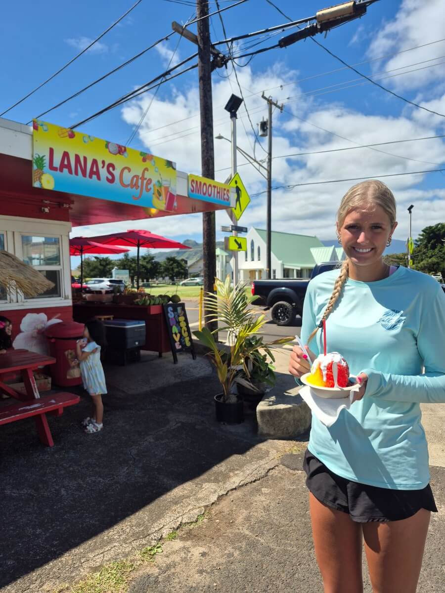 A girl smiles in front of a sign stating Lana's Cafe holding a red and yellow shaved ice