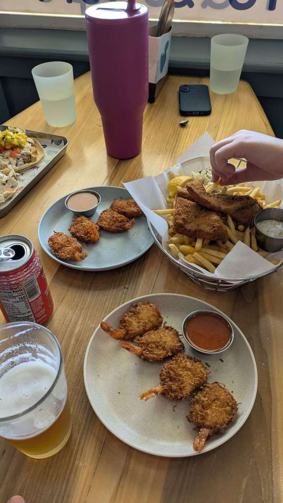 A spread of fish and chips, coconut shrimp, and tacos set on a table among various drinks.