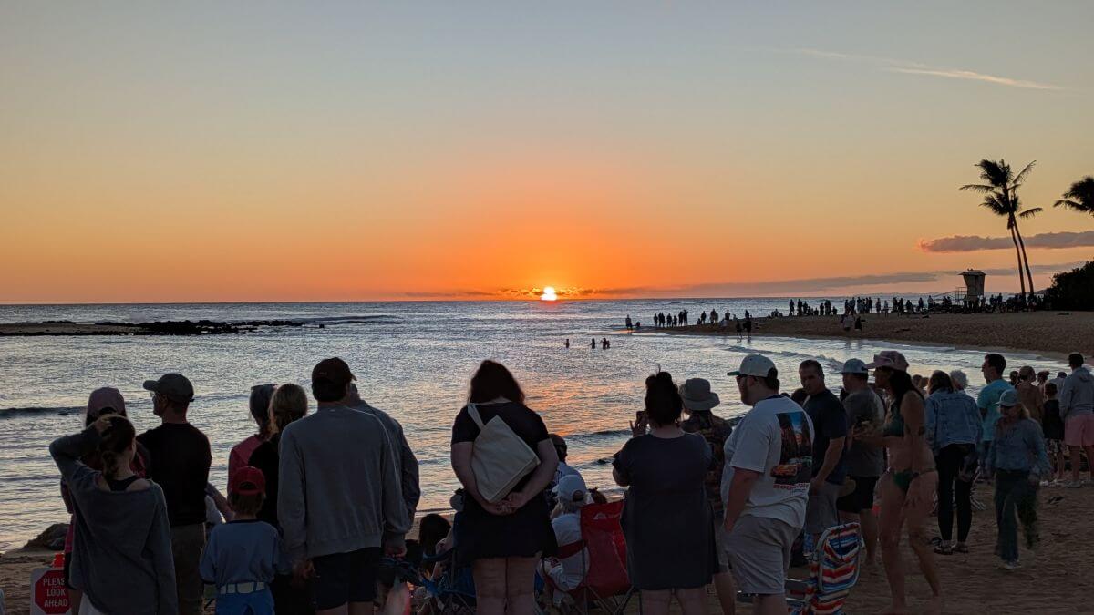 The silhouette of a crowd watching sea turtles rest on a beach trims out the ocean as a yellow sun sinks below the silver ocean, the sky fading from orange to pale blue.