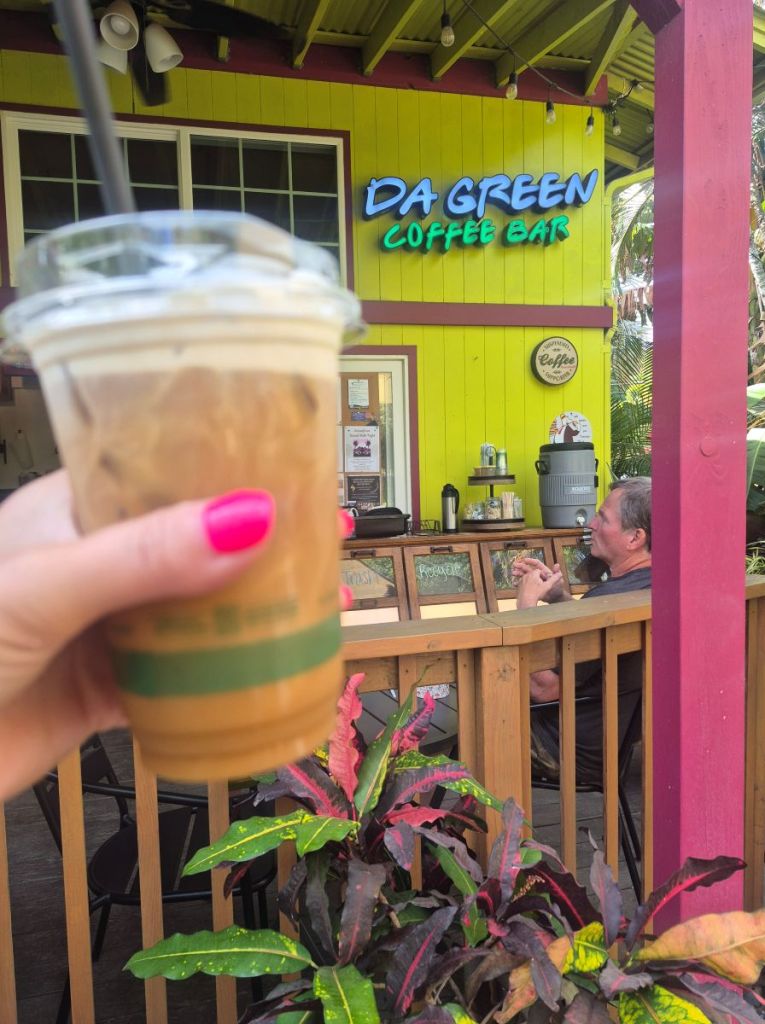 An iced coffee held up in front of a yellow building with an illuminated Da Green Coffee Bar sign