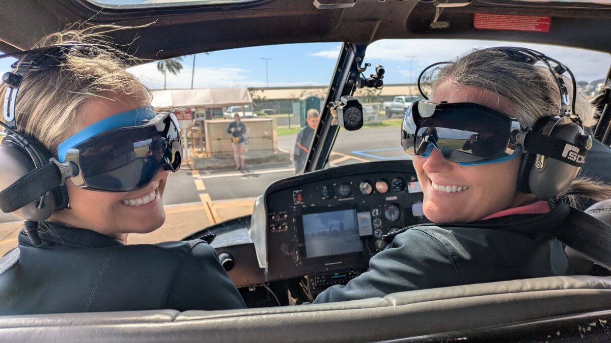 Two girls smile at the camera in the front seat of a helicopter wearing goggles and large headphones