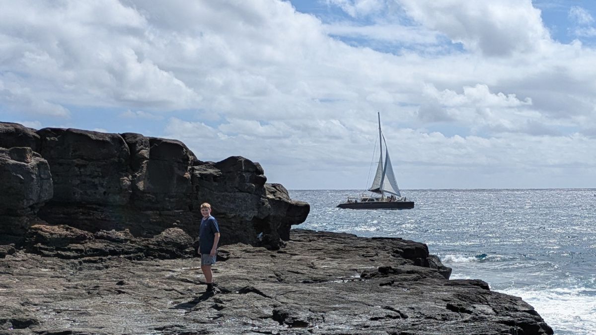 A boy stands on black lava rock as a sailboat passes by in the distance.