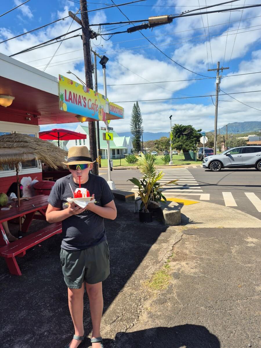 A boy in gray slurps from a bowl of shaved ice in a brown fedora
