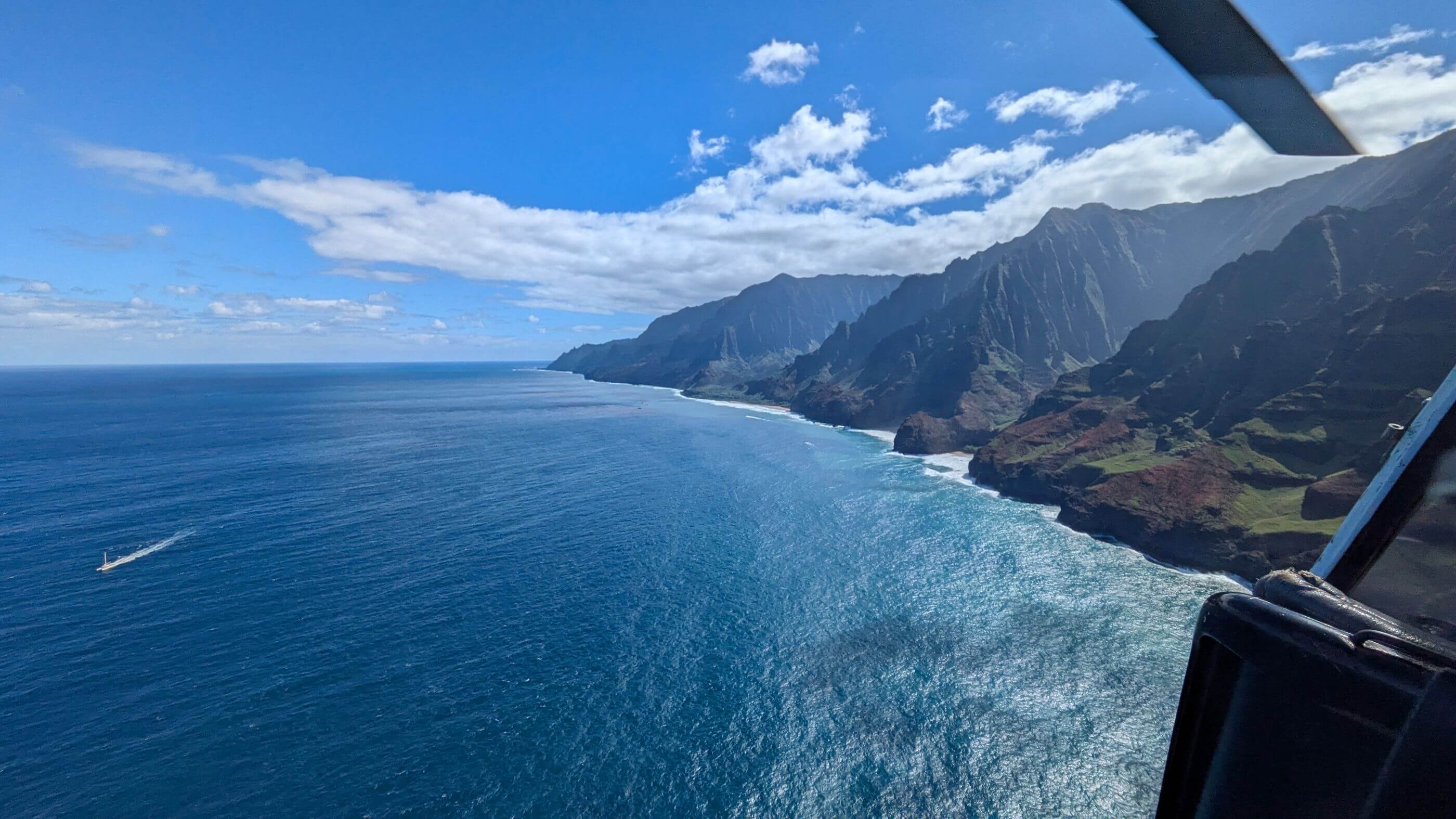 Gorgeous green mountains tower over the deep blue Pacific as a boat cruises along with a long white tail following