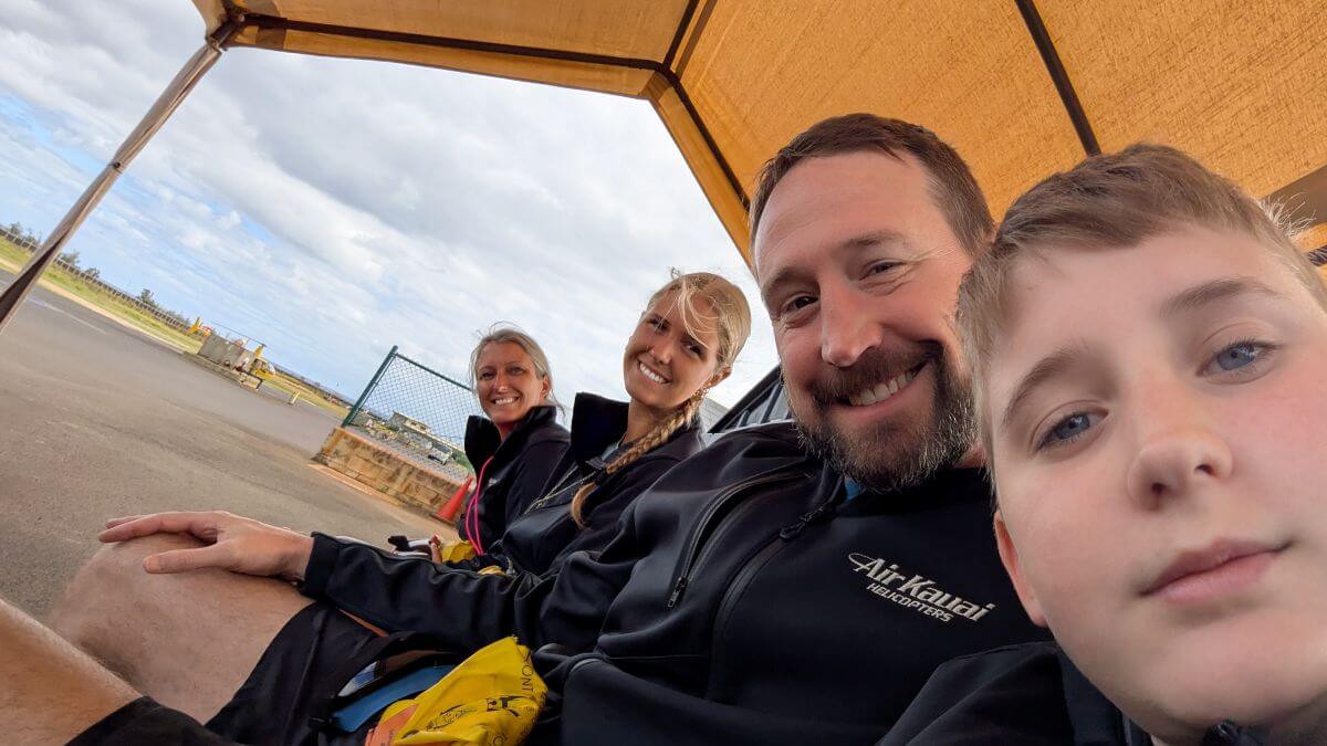 A family of four sits under a tent at the airport next to a helicopter landing pad