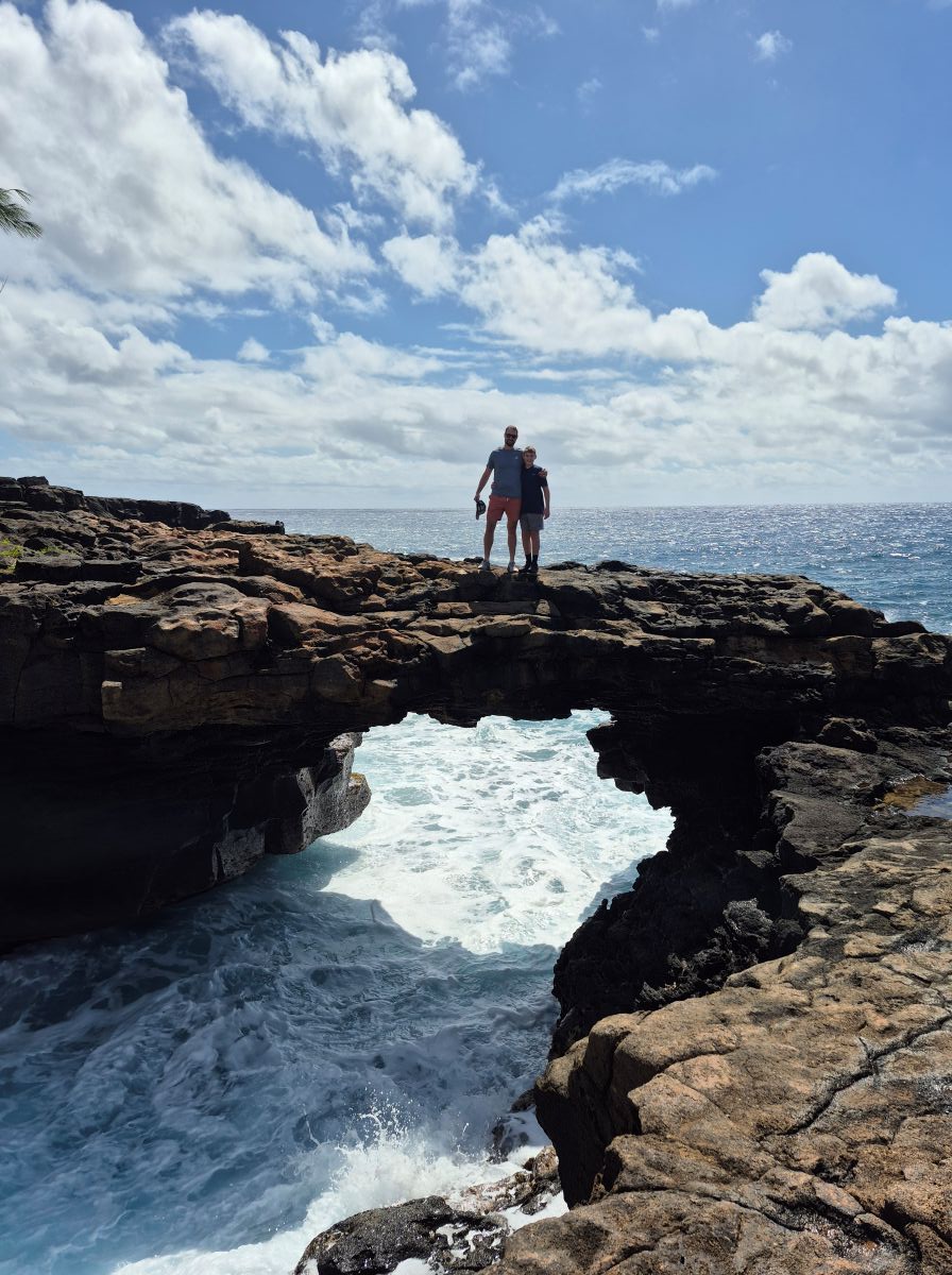 A father and son stand on a natural arch over the foamy ocean.