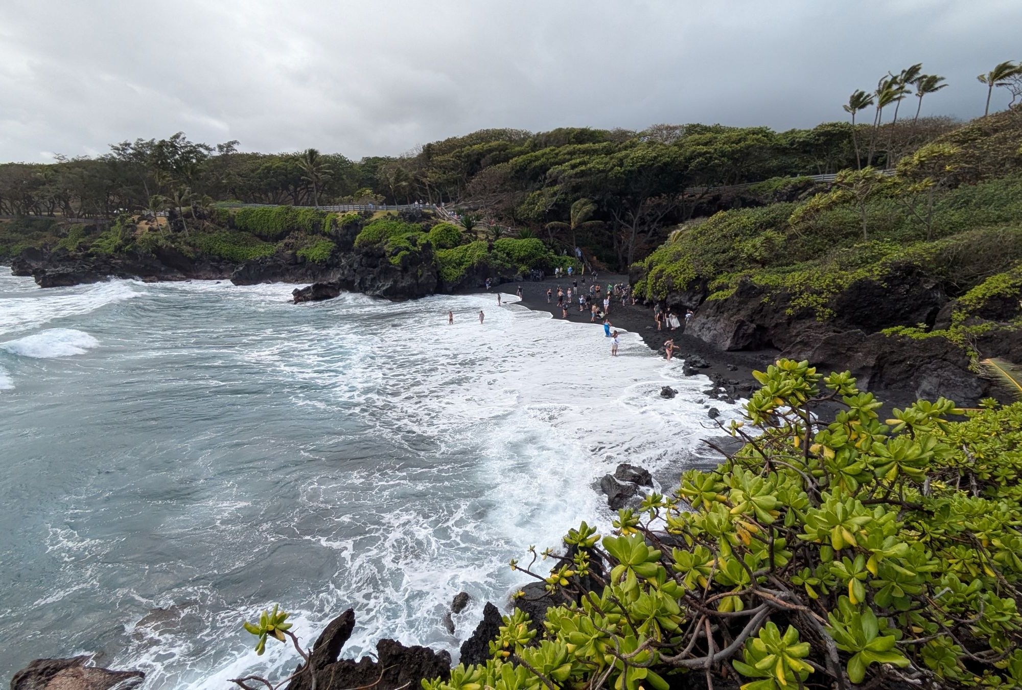 Waianapanapa State Park from the West. A black sand beach with people scattered around.