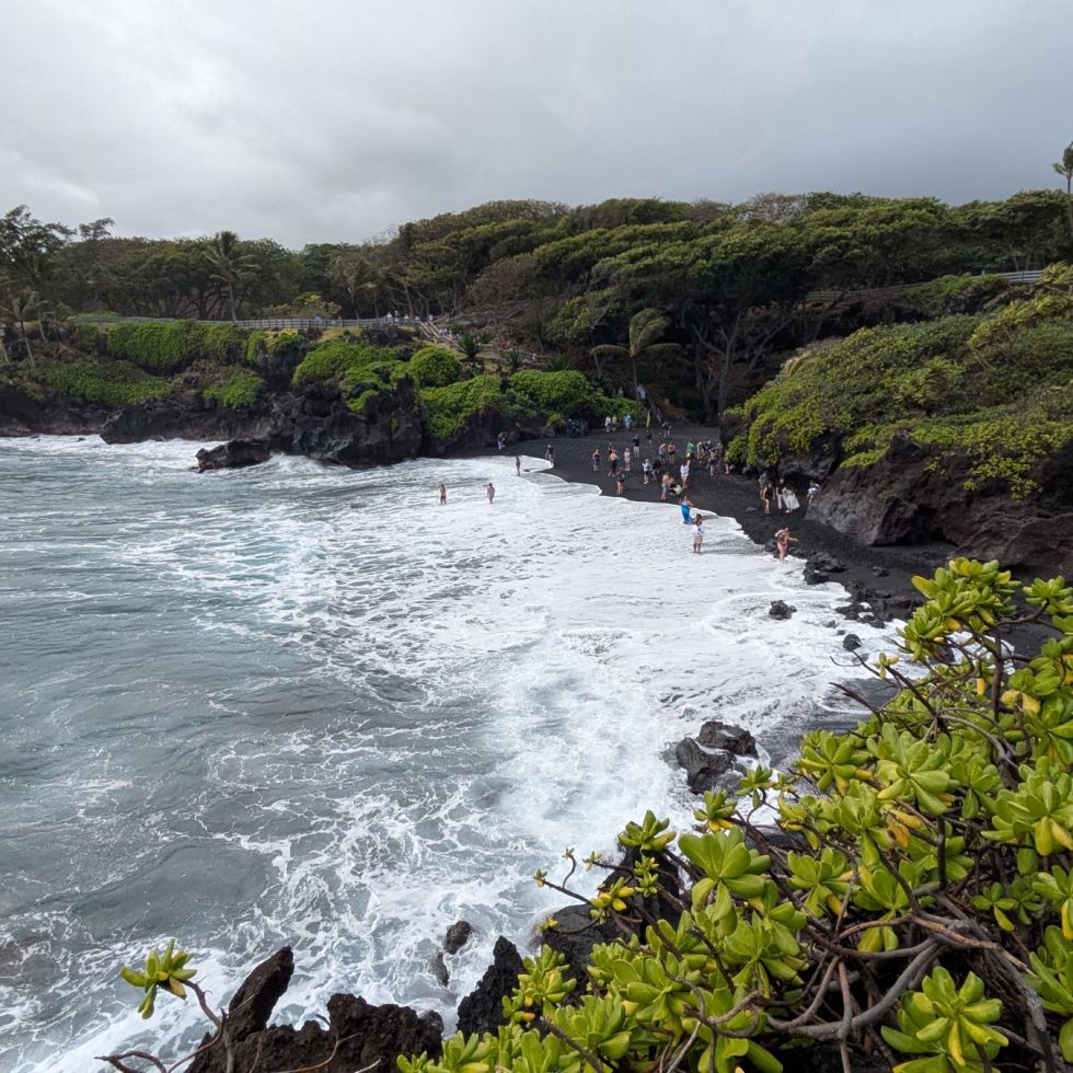 Waianapanapa State Park from the West. A black sand beach with people scattered around.